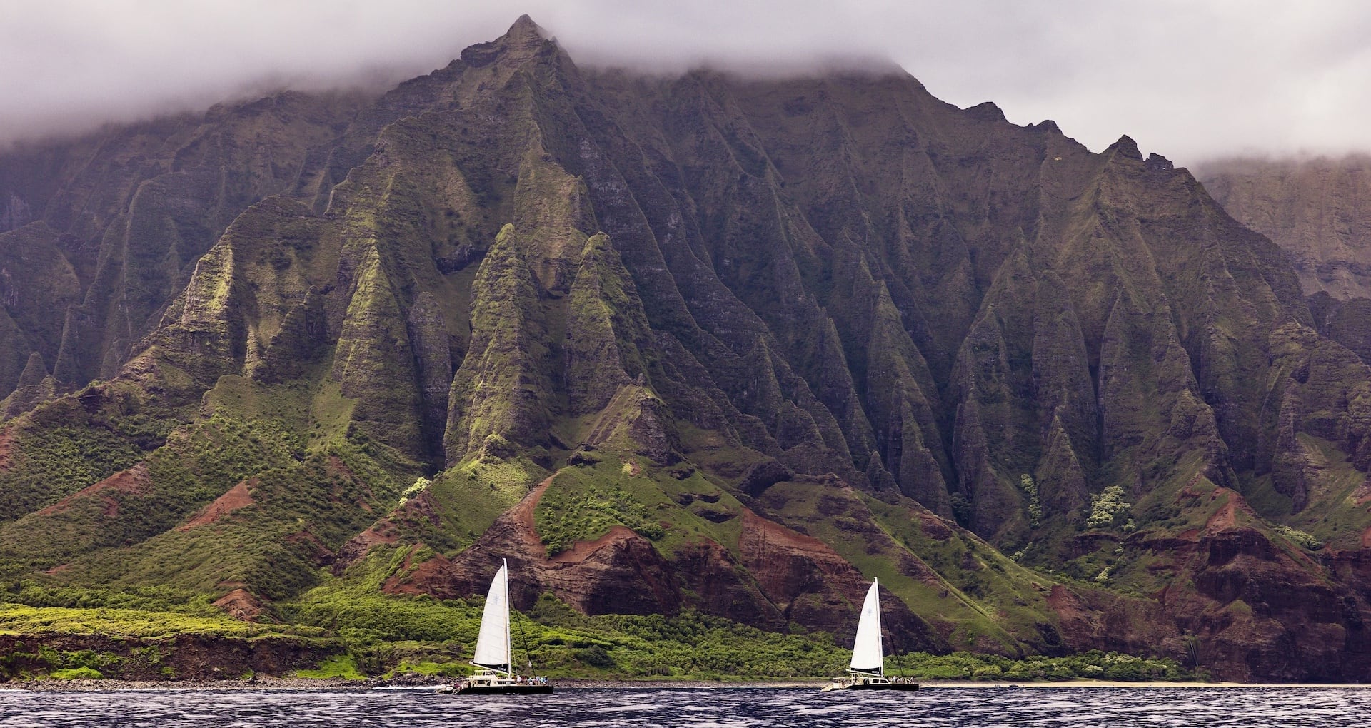 Two sailboats gliding along the base of the towering green cliffs of the Na Pali Coast