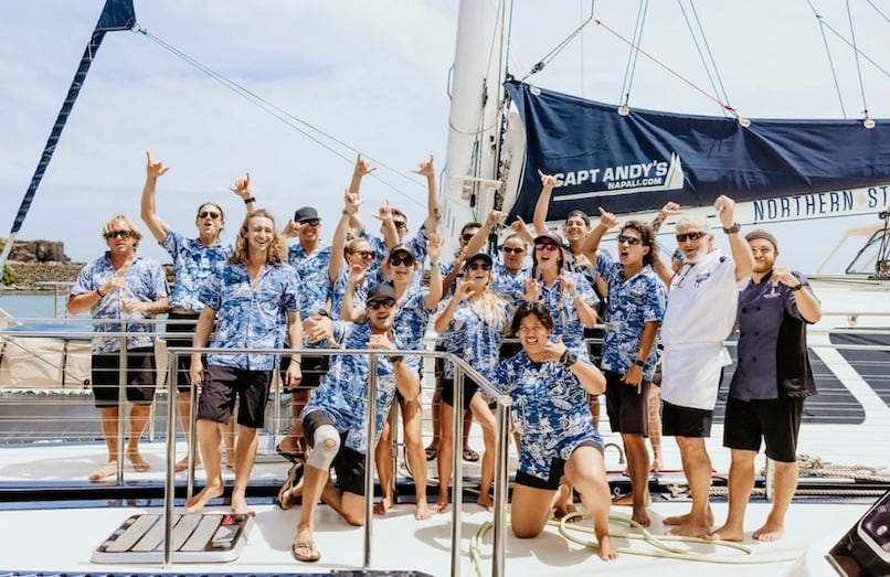 Crew of Captain Andy's posing in blue Hawaiian shirts on a boat, making shaka signs with the Captain Andy's banner visible in the background.