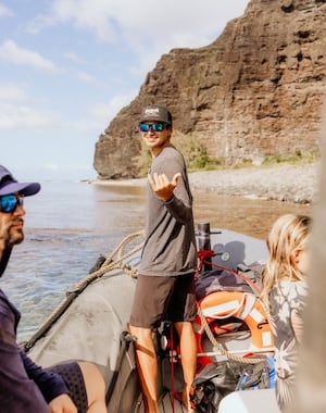 Captain Andy's crew member smiling and making a shaka sign on a boat near the Na Pali Coast, with rocky cliffs and the ocean in the background.
