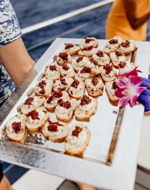 Tray of appetizers with an orchid garnish being served on a boat, with the ocean and guests in the background.