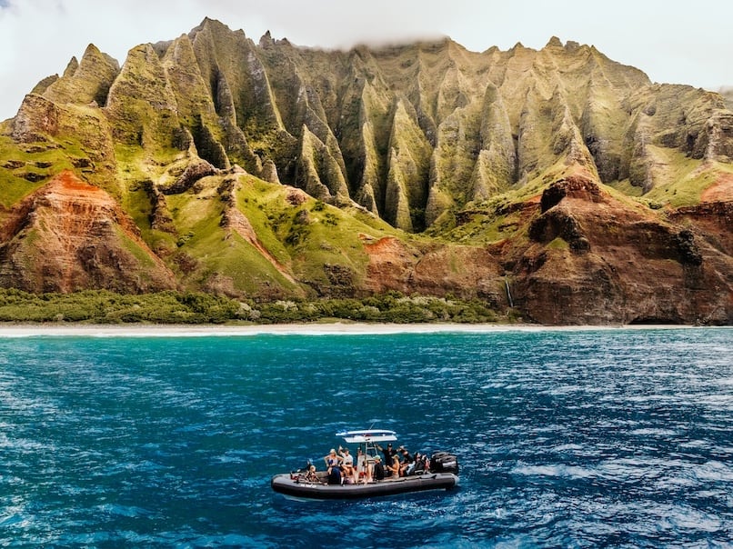 A boat full of people floating near the lush, green cliffs of the Na Pali Coast