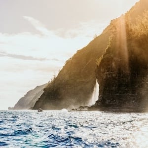 Sunlight streaming over the cliffs of the Na Pali Coast, with ocean waves crashing against the rocky shoreline
