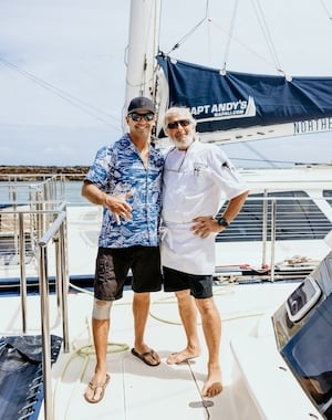 Captain Andy's crew members posing on a sailboat, with one wearing a blue Hawaiian shirt and the other in a white uniform, both making shaka signs.