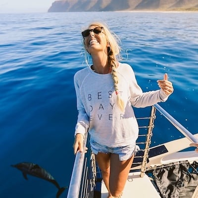 Woman smiling and making a shaka sign on a boat, with dolphins swimming in the background.