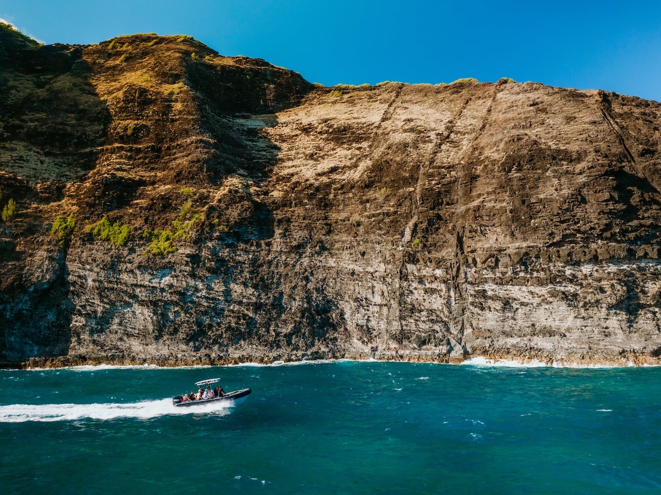 A boat speeding along the base of towering rocky cliffs of the Na Pali Coast, with clear blue ocean water and a bright sky.