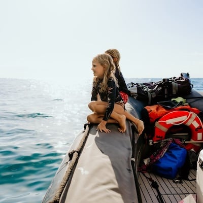 Two children sitting on the edge of a boat, looking out at the ocean and enjoying the view, with life jackets and gear visible on the boat.