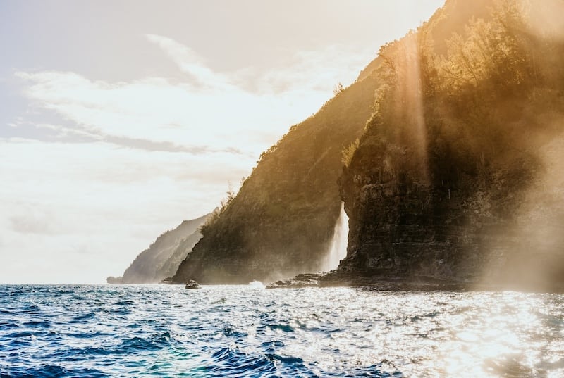 Sunlight streaming over the cliffs of the Na Pali Coast, with ocean waves crashing against the rocky shoreline.