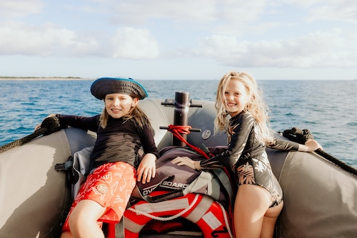Two smiling children sitting on the edge of a boat, enjoying the ocean view on a sunny day.