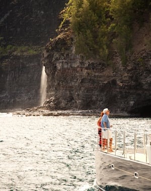 Couple standing on the deck of Captain Andy's boat, admiring a waterfall cascading down the rocky cliffs of the Na Pali Coast.