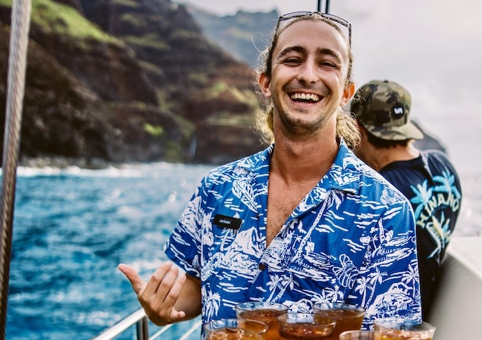 A smiling crew member from Captain Andy's offers refreshments aboard a boat with the Na Pali Coast in the background.