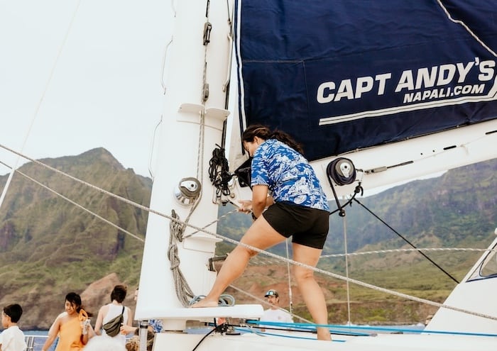 A crew member climbing the rigging on a Captain Andy's sailboat, with lush green mountains of the Na Pali Coast in the background.