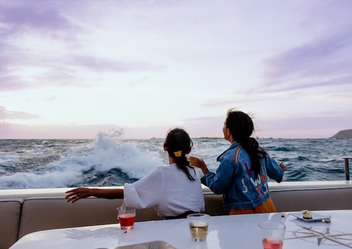 Two women enjoying a sunset view from a boat on the ocean, with drinks on the table and waves crashing nearby.