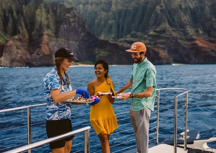 A sandwich, pineapple slices, and a can of Aloha Strawberry Orange drink on a table with two people sitting by the ocean in the background.