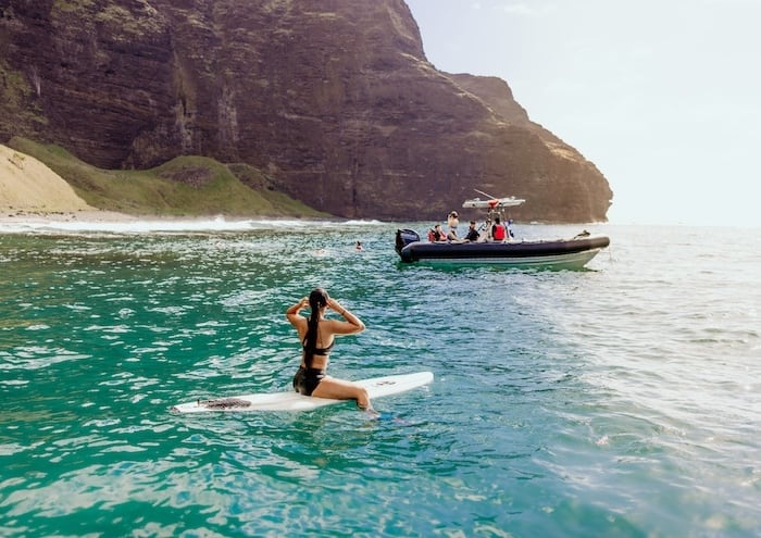 A woman on a paddleboard in the ocean near the rocky cliffs of the Na Pali Coast, with a boat full of people in the distance.