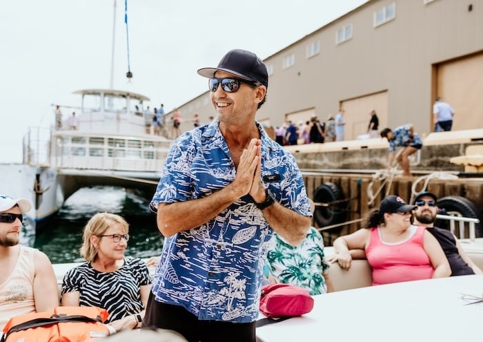 A smiling crew member of Captain Andy's in a blue Hawaiian shirt greeting passengers near a docked boat, with passengers sitting and preparing for a tour.