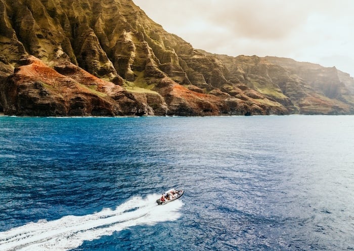 Boat speeding along the Na Pali Coast, showcasing the dramatic, green and red cliffs and the expansive blue ocean.